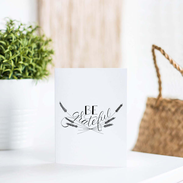 Be Grateful A2 Greeting Card, black hand lettered design with wheat illustrations on a folded white card shown on a table with a plant and handbag