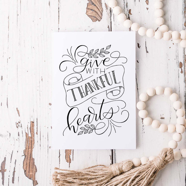Give with thankful hearts hand lettered greeting card design in black on a folded white card with A2 envelope shown on white rustic wooden table with white wooden bead garland