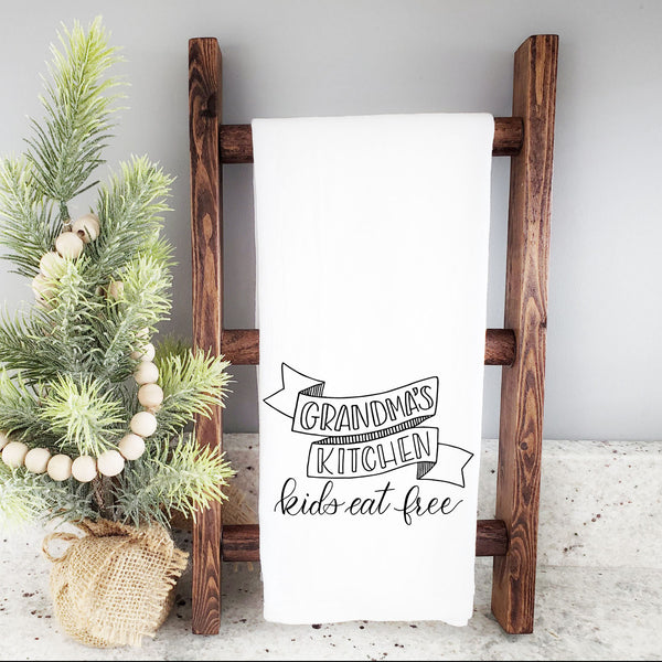 White floursack kitchen towel with black hand lettered illustration that says grandma's kitchen kids eat free shown hanging folded from a wooden display ladder with a mini Christmas tree
