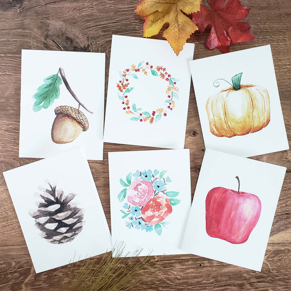 Set of 6 Watercolor painted notecards in fall autumn nature designs including an acorn with leaf, orange pumpkin, fall foliage wreath, red apple, fall floral bouquet and pinecone shown on a wooden table