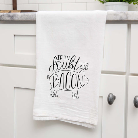 White floursack kitchen towel with black hand lettered illustrated design that says If in doubt add bacon with a outline of a pig shown folded and hanging from a countertop in a modern kitchen