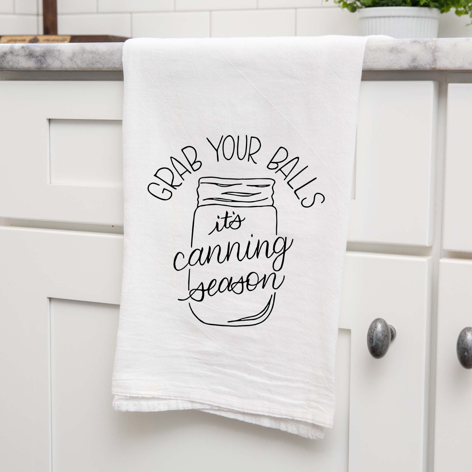 White floursack kitchen towel with black hand lettered illustrated design that says Grab your balls it's canning season with a mason jar doodle shown folded and hanging from a countertop in a modern kitchen