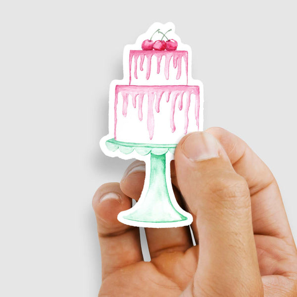 3" vinyl sticker of a watercolor painted 2-tiered white birthday cake with pink frosting and 3 cherries on top all sitting on a vintage scalloped mint colored cake stand shown with sticker being held by a woman's hand