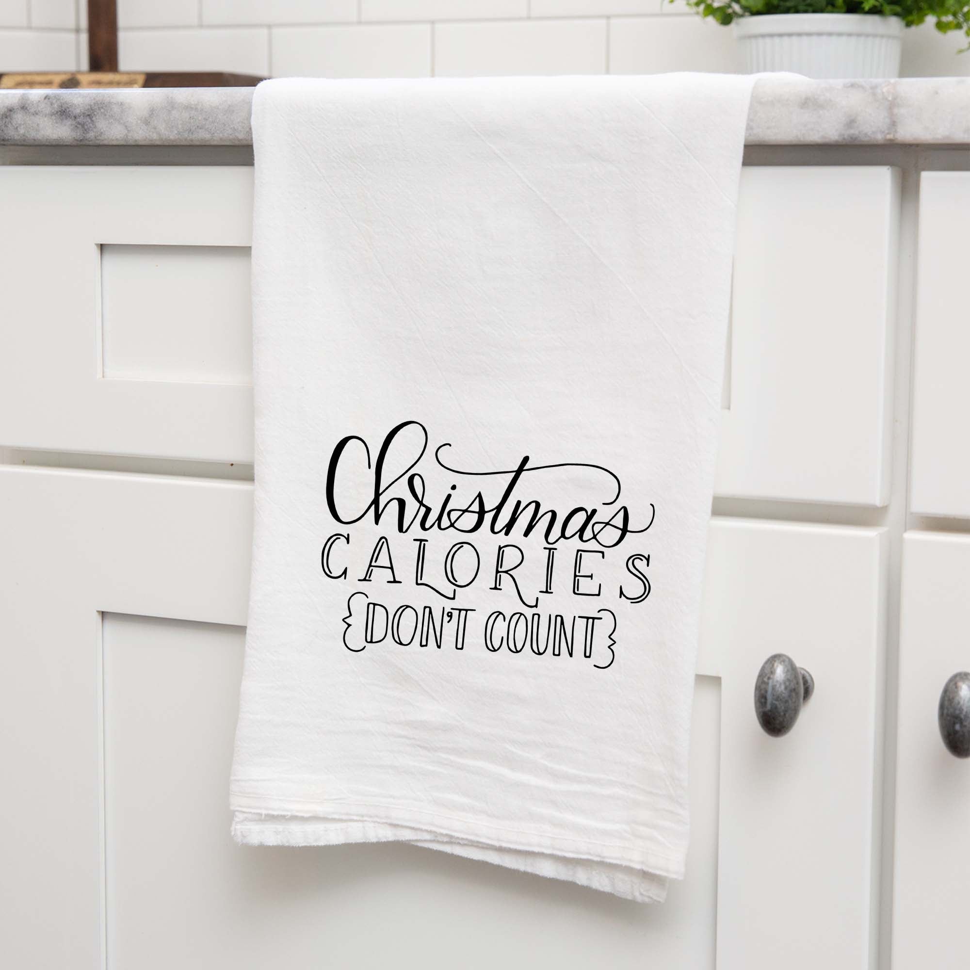 White floursack towel with black hand lettered illustrated design that says Christmas calories don't count shown folded and hanging from a countertop in a modern kitchen