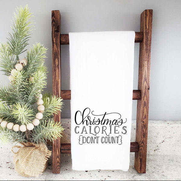 White floursack towel with black hand lettered illustrated design that says Christmas calories don't count shown folded and hanging from a wooden display ladder and mini Christmas tree