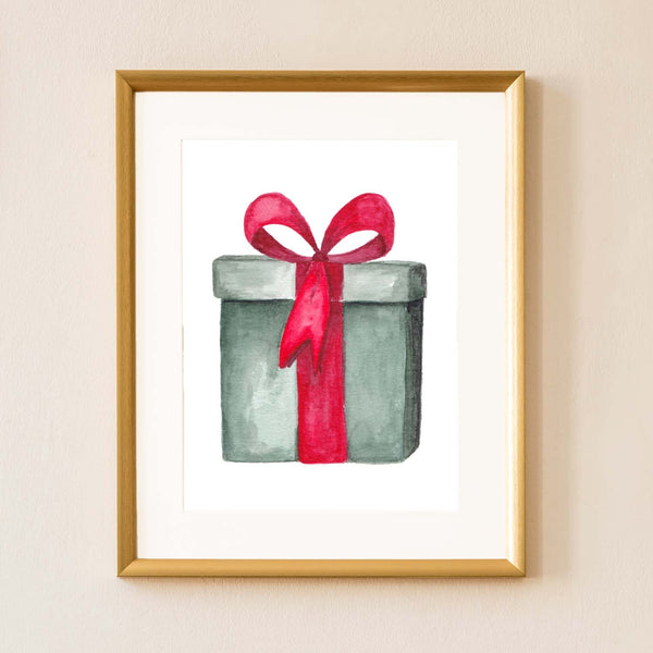 Watercolor painting of a green christmas package tied with a red bow shown hanging on the wall in a gold frame