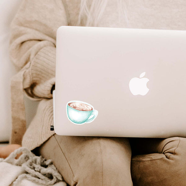 3" vinyl sticker of a watercolor blue coffee mug full of hot coffee shown adhered to a MacBook laptop cover sitting open on a woman's lap