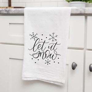 White Floursack Kitchen Towel with black hand lettered illustration that says let it snow with snowflake doodles shown hanging from a countertop in a modern kitchen