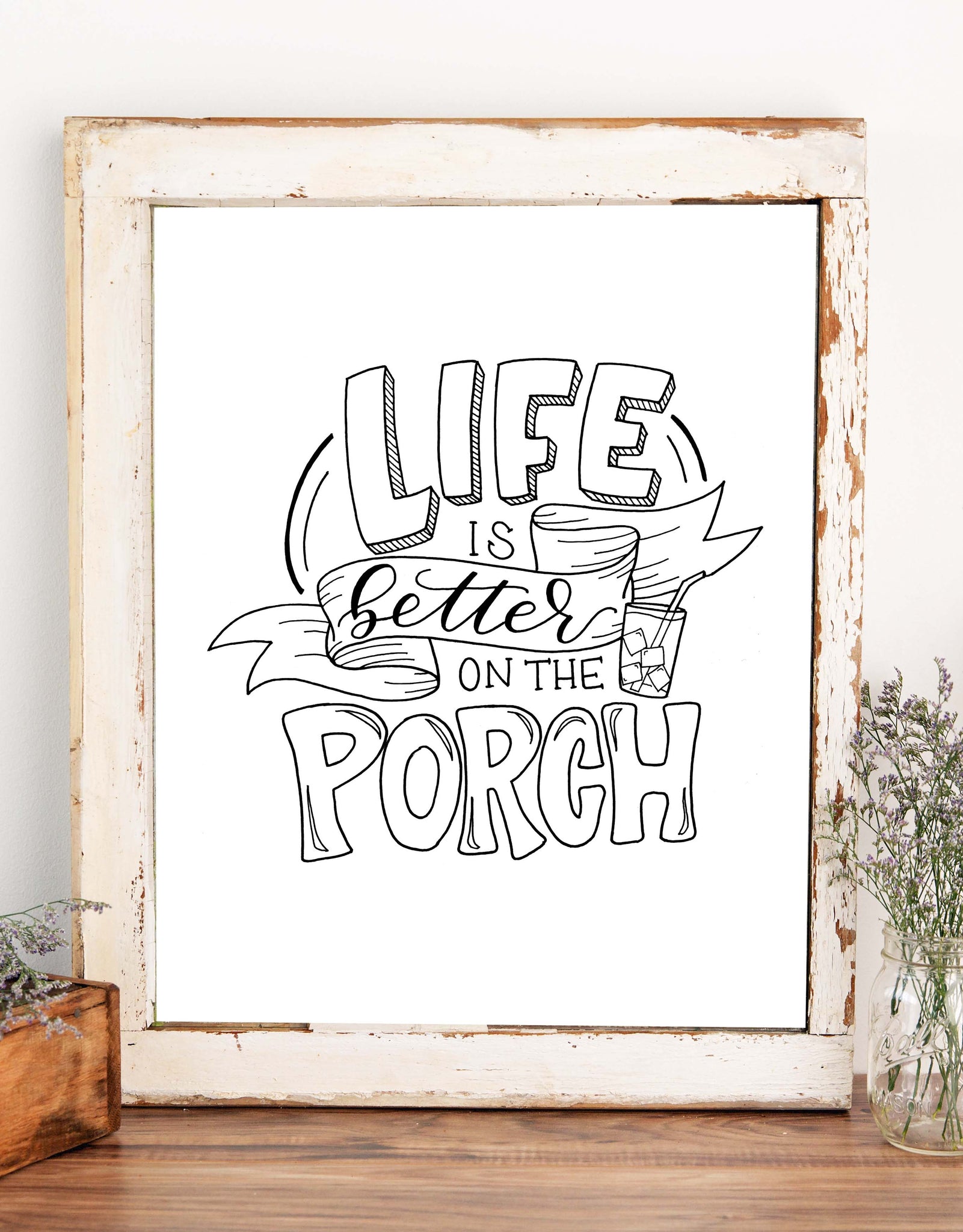 Hand lettered illustrated wall art design that says life is better on the porch with an illustrated banner and drink in a glass of ice with a straw shown in a rustic white wooden frame with wispy flowers in a box and jar