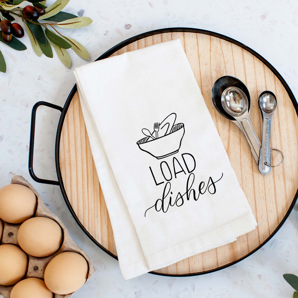 White floursack towel with black hand lettered illustrated design that says Load dishes with pile of dirty dishes doodles shown folded on a serving tray with a set of measuring spoons and fresh eggs