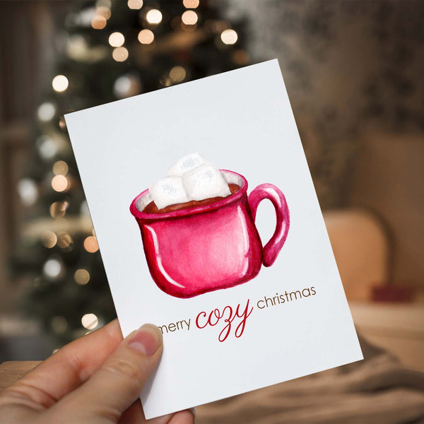 Watercolor Christmas greeting card with a watercolor painted red mug full of hot cocoa and heaping with marshmallows that says merry cozy christmas  shown with a woman holding card in front of a lighted christmas tree