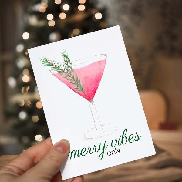 Watercolor greeting card with a watercolor christmas cocktail that says merry vibes only shown with a woman holding card in front of a lighted christmas tree