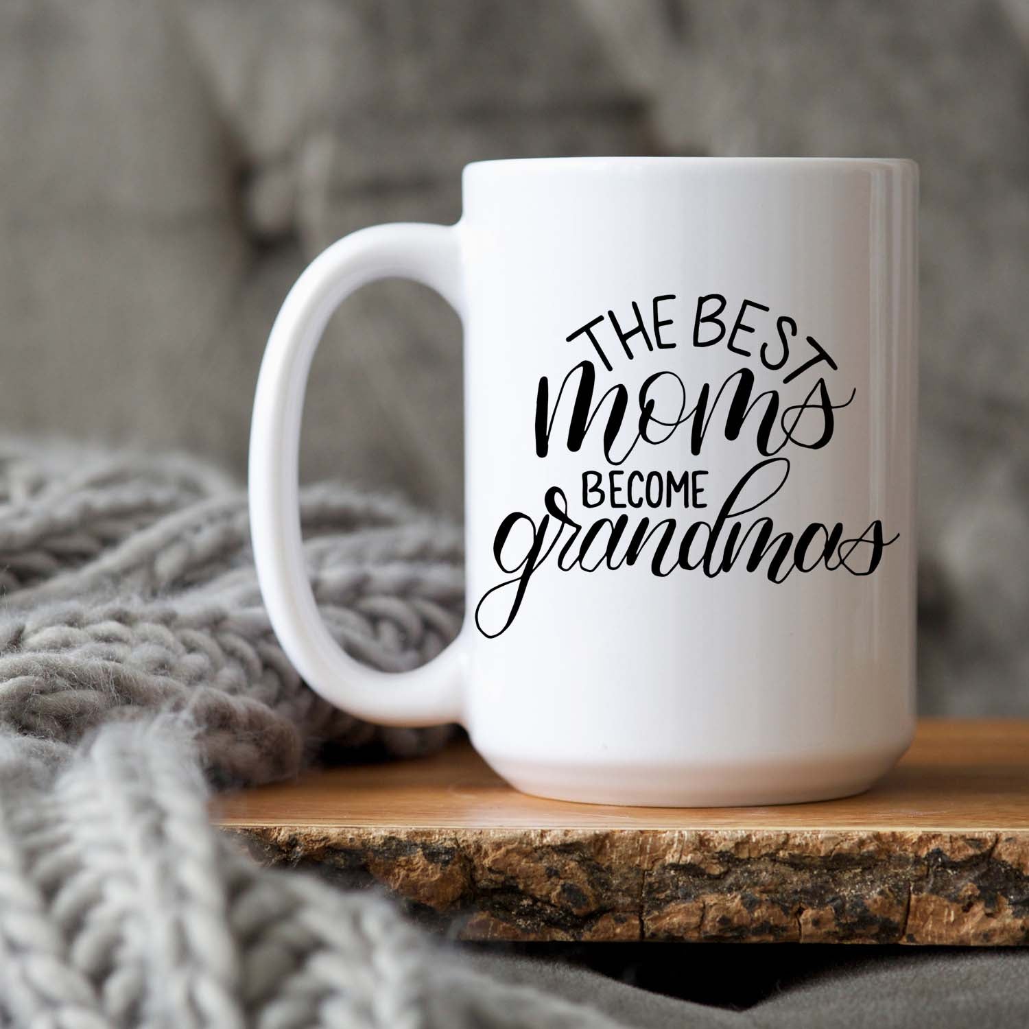 15oz white ceramic mug with hand lettered illustrated design that says the best moms become grandmas shown sitting on a wood tray with a grey knit blanket