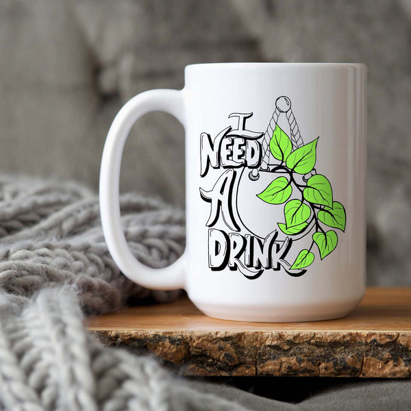 15oz white ceramic mug with hand lettered illustrated design that says I need a drink with an illustration of a pothos plant with green leaves in a hanging pot shown sitting on a wood tray with a grey knit blanket