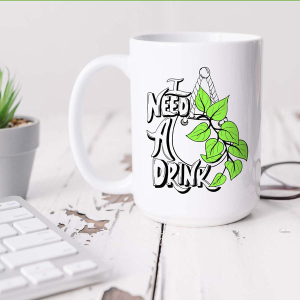 15oz white ceramic mug with hand lettered illustrated design that says I need a drink with an illustration of a pothos plant with green leaves in a hanging pot shown sitting on a white office desk