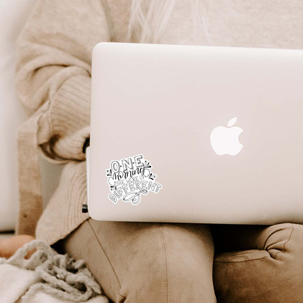 3" hand lettered, illustrated, black and white vinyl sticker that says one morning she woke up different shown adhered to a MacBook laptop cover sitting open on a woman's lap