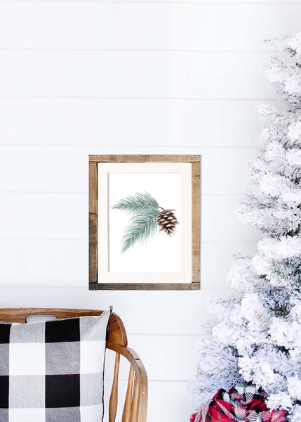 Watercolor painting of evergreen branches and pinecone shown in a wood frame hanging on a shiplap wall next to a white flocked tree and rocking chair