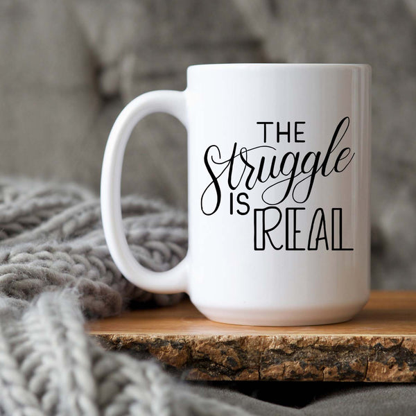 15oz white ceramic mug with hand lettered illustrated design that says the struggle is real shown sitting on a wood tray with a grey knit blanket