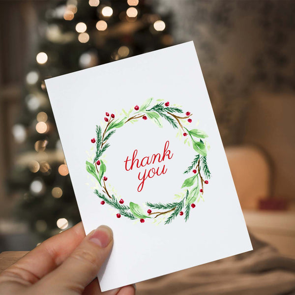 Watercolor Christmas greeting card with a holiday wreath of twigs, greens and berries in red and green that says thank you shown with a woman holding in front a lighted christmas tree